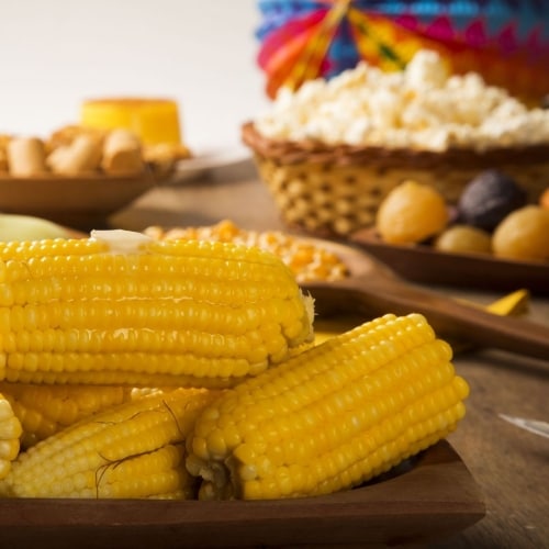 Mesa de comidas juninas com destaque para espigas de milho cozido + pipoca, doce de abóbora e paçoca ao fundo
