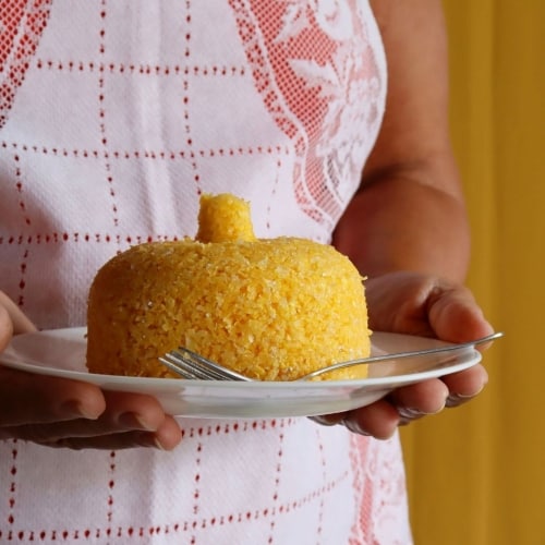 Mulher usando avental, segurando prato branco com cuscuz nordestino e garfo