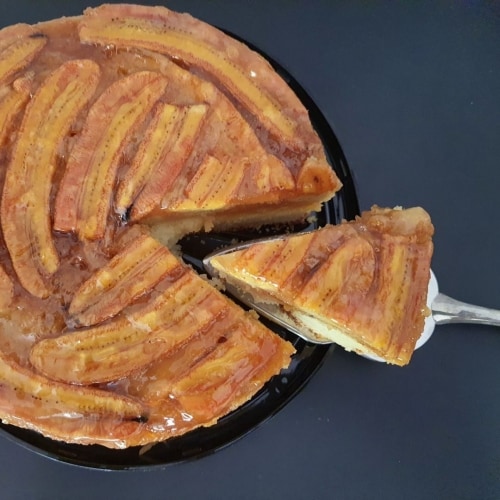 Bolo rústico de banana, visto de cima, com espátula cortando um pedaço triangular