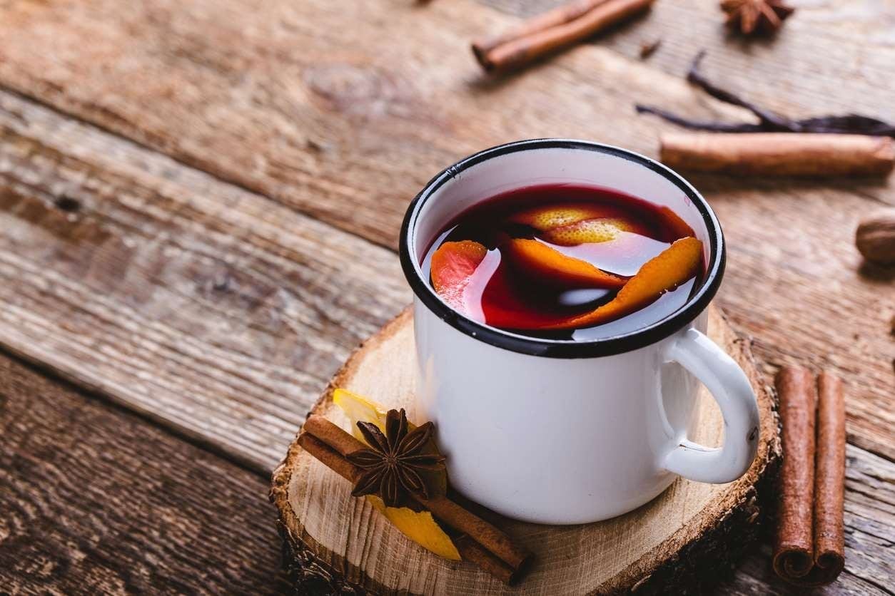 Caneca de ágata com vinho quente e casca de laranja, sobre mesa de madeira, com aniz-estrelado e paus de canela ao redor