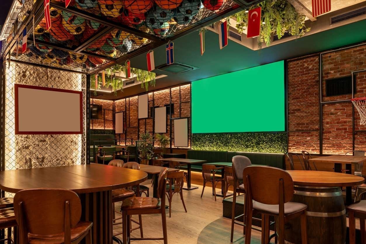 Interior de bar com temática esportiva, bolas e bandeiras fazendo referência à Copa do Mundo, além de um telão ao fundo.