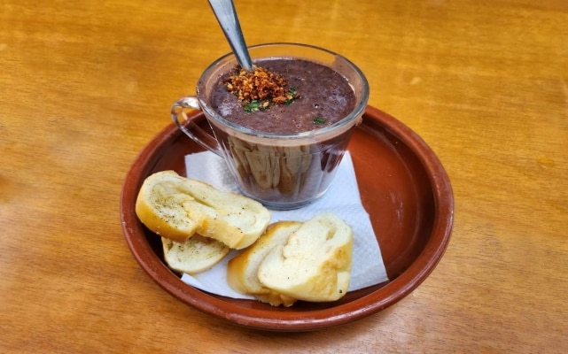 Porção de caldinho de feijão preto, servido com farias de pão francês e crocante de bacon e alho. Porção de caldinho de feijão preto, servido com farias de pão francês e crocante de bacon e alho.
