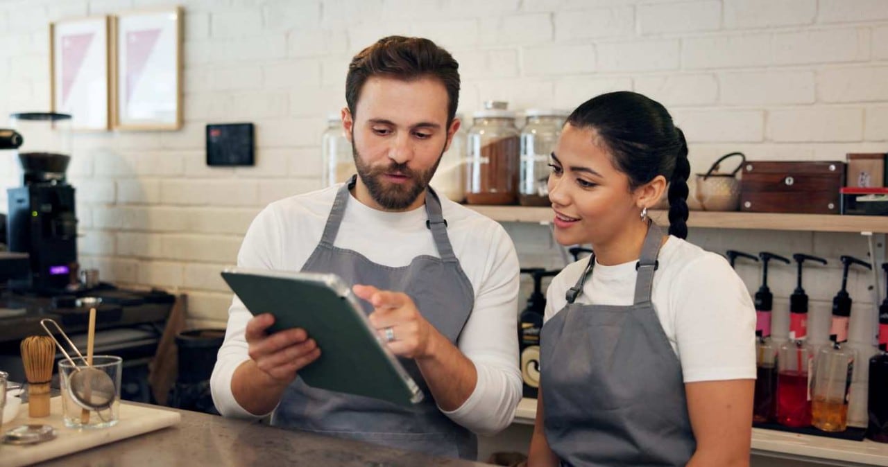Dois colaboradores conversam em ambiente de cozinha, lendo algo juntos em um tablet