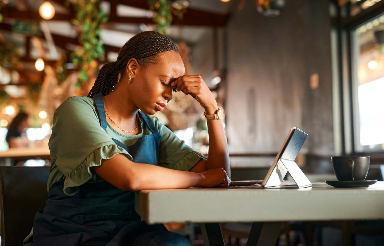 Dona de cafeteria parece muito estressada, olhos fechados, mãos no rosto, frente a um tablet