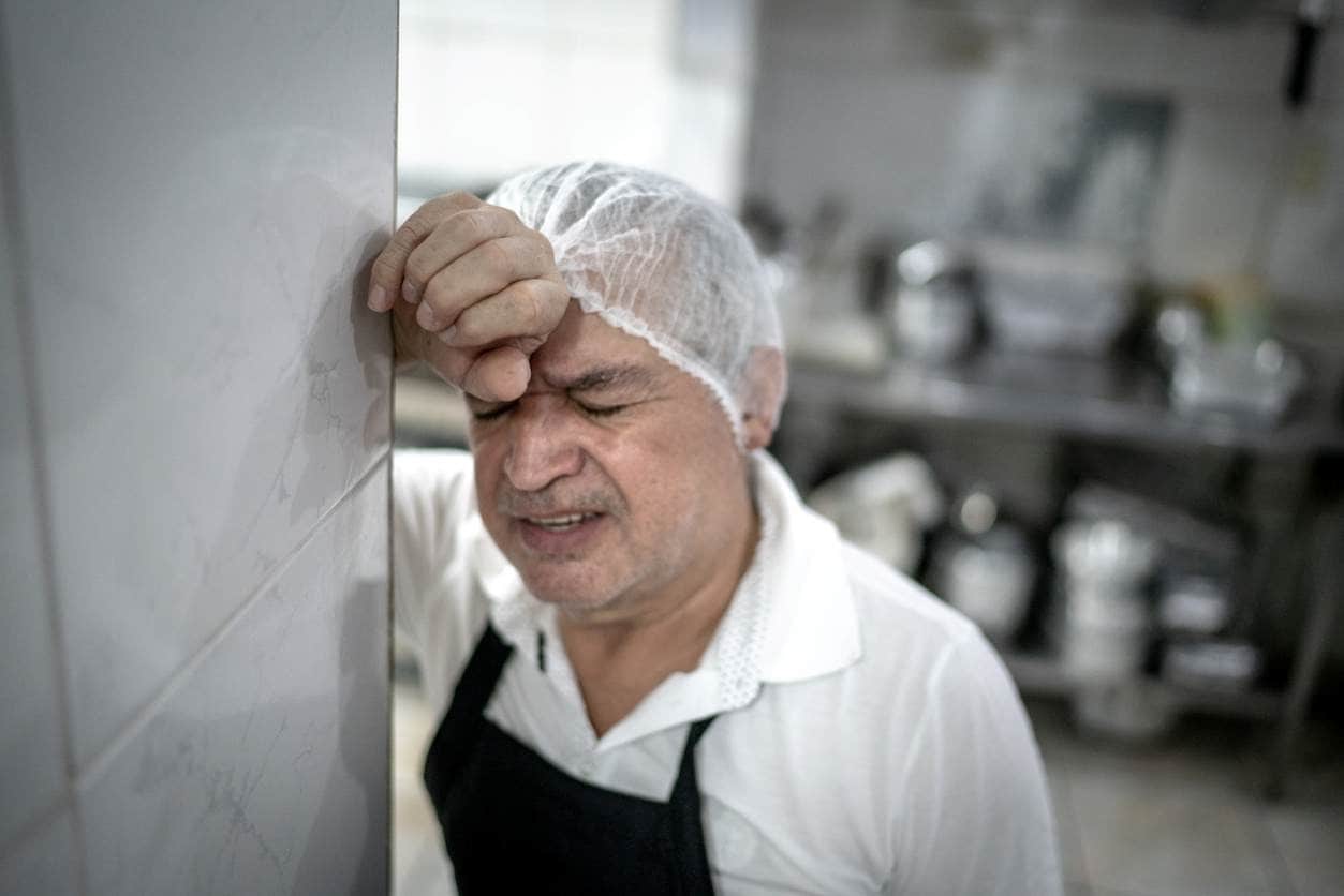 Ajudante de cozinha, com semblante preocupado, encostado na parede do seu ambiente de trabalho