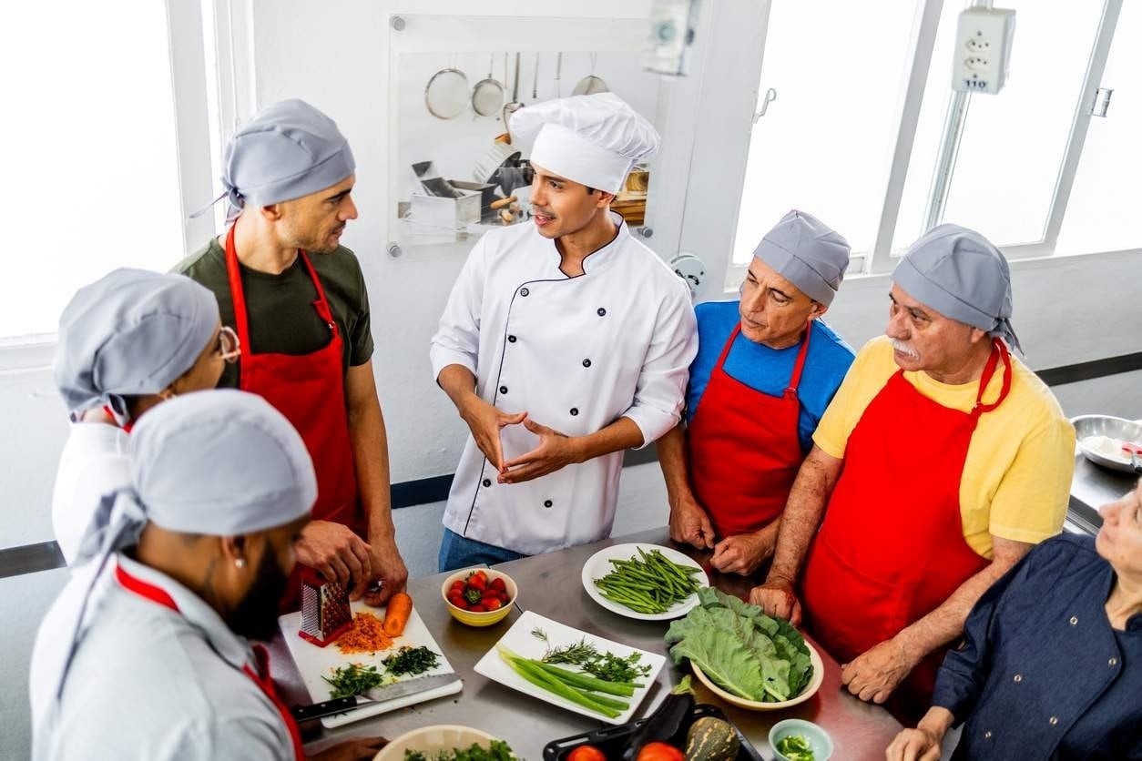 Chef de cozinha dando aula sobre preparação de alimentos e conversando com a equipe.