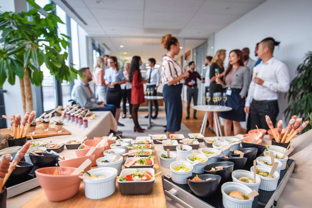 Coffee break corporativo muito bem servido, com pessoas ao fundo conversando e bebendo espumante