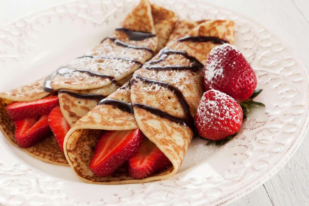 Panquecas doces enroladas com morangos frescos, calda de chocolate e açúcar de confeiteiro em prato branco.