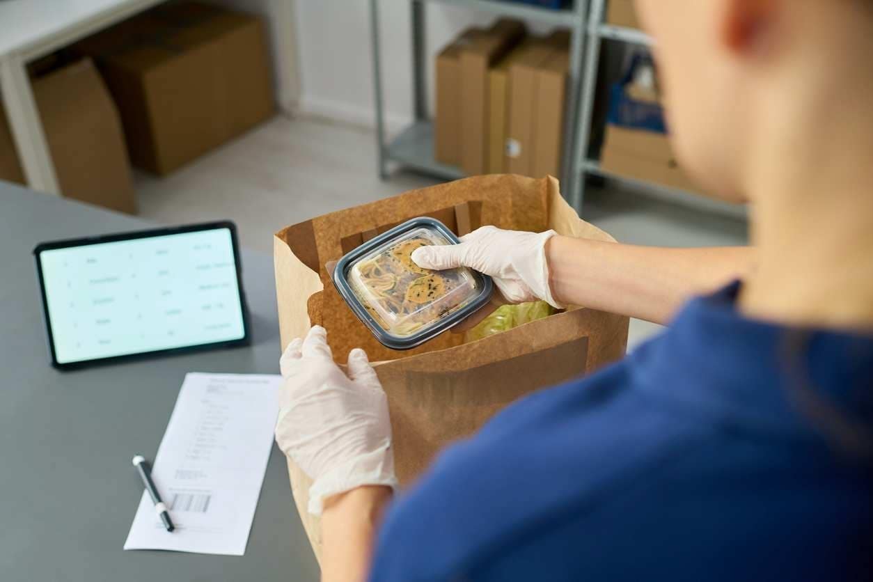 Pessoa com luvas embala marmita em sacola de papel para delivery, com tablet de pedidos ao lado, ilustrando a operação de uma loja no iFood.