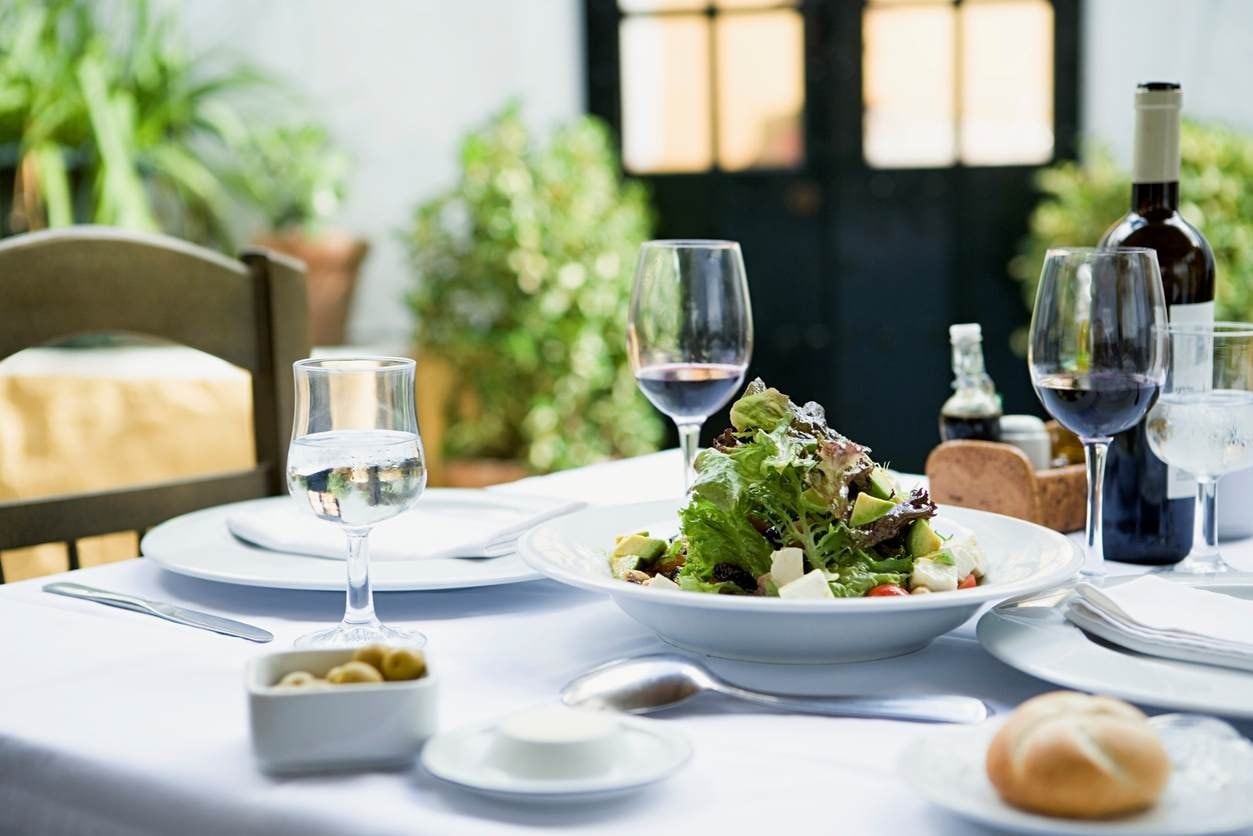 Mesa de jantar posta, com diversos pratos, talheres, vinho e taças. Destaque para uma saladinha ao centro.