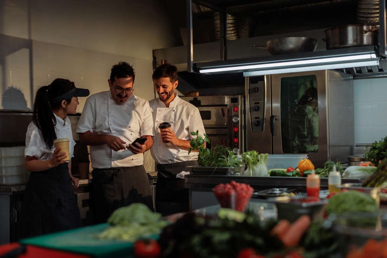 Cozinheiros e chef conversando, vendo algo no celular juntos e tomando um café na cozinha.