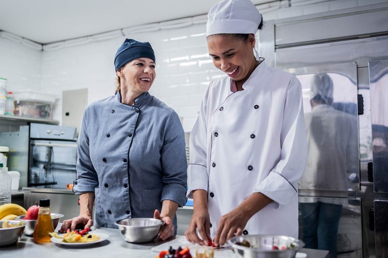 Duas profissionais de cozinha conversando e sorrindo enquanto preparam prato doce.