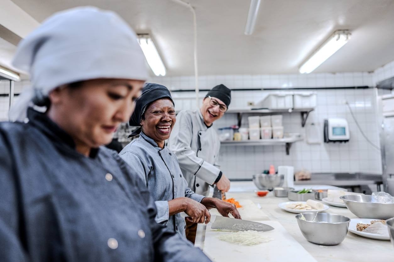 Pessoas picando ingredientes em cozinha profissional, usando vestes adequadas e sorrindo para a foto. Pessoas picando ingredientes em cozinha profissional, usando vestes adequadas e sorrindo para a foto.