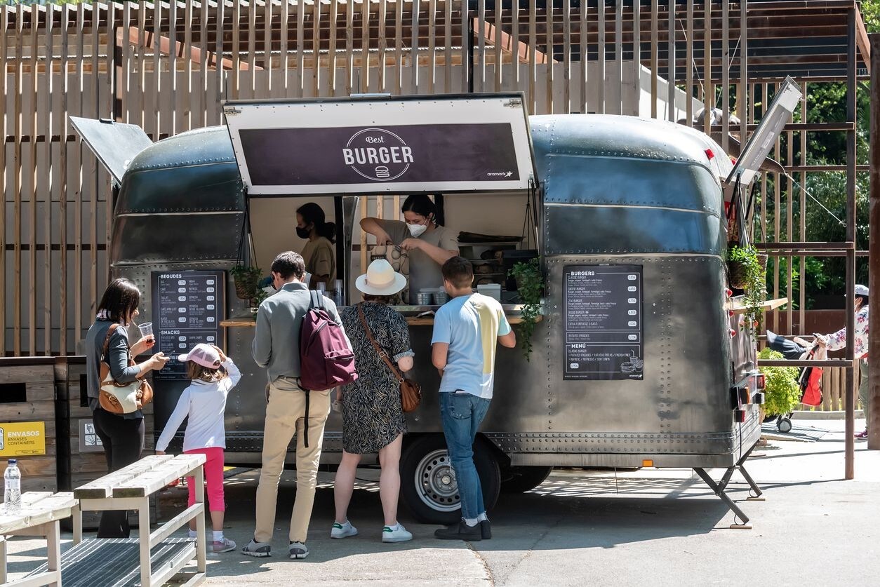 Trailer de comida estacionado em praça apropriada, com duas funcionárias atendendo fila de clientes. Trailer de comida estacionado em praça apropriada, com duas funcionárias atendendo fila de clientes.