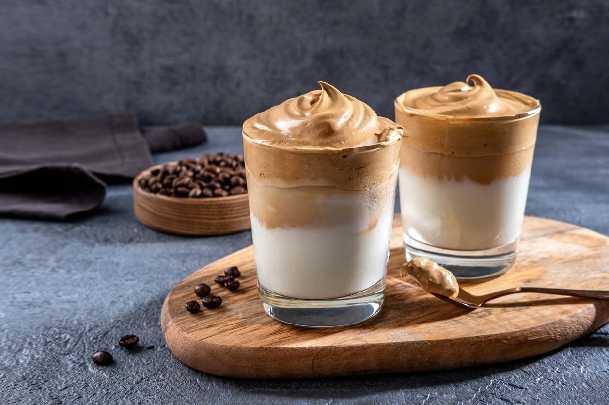 Dois copos com Dalgona Coffee, servidos sobre tábua de madeira, com grãos de café espalhados como decoração.