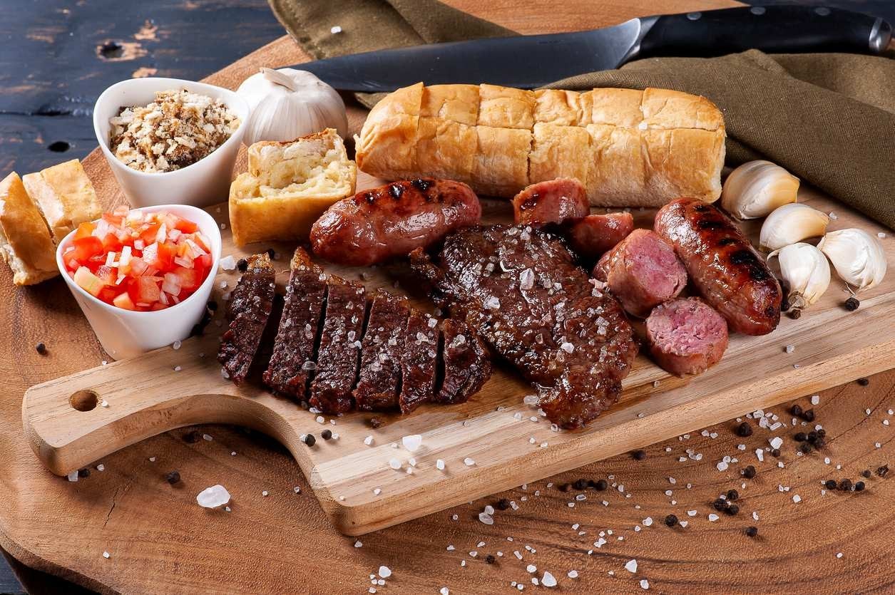 Churrasco típico brasileiro, com linguiça, picanha, pão de alho, vinagrete e farofa, sobre tábua de madeira.