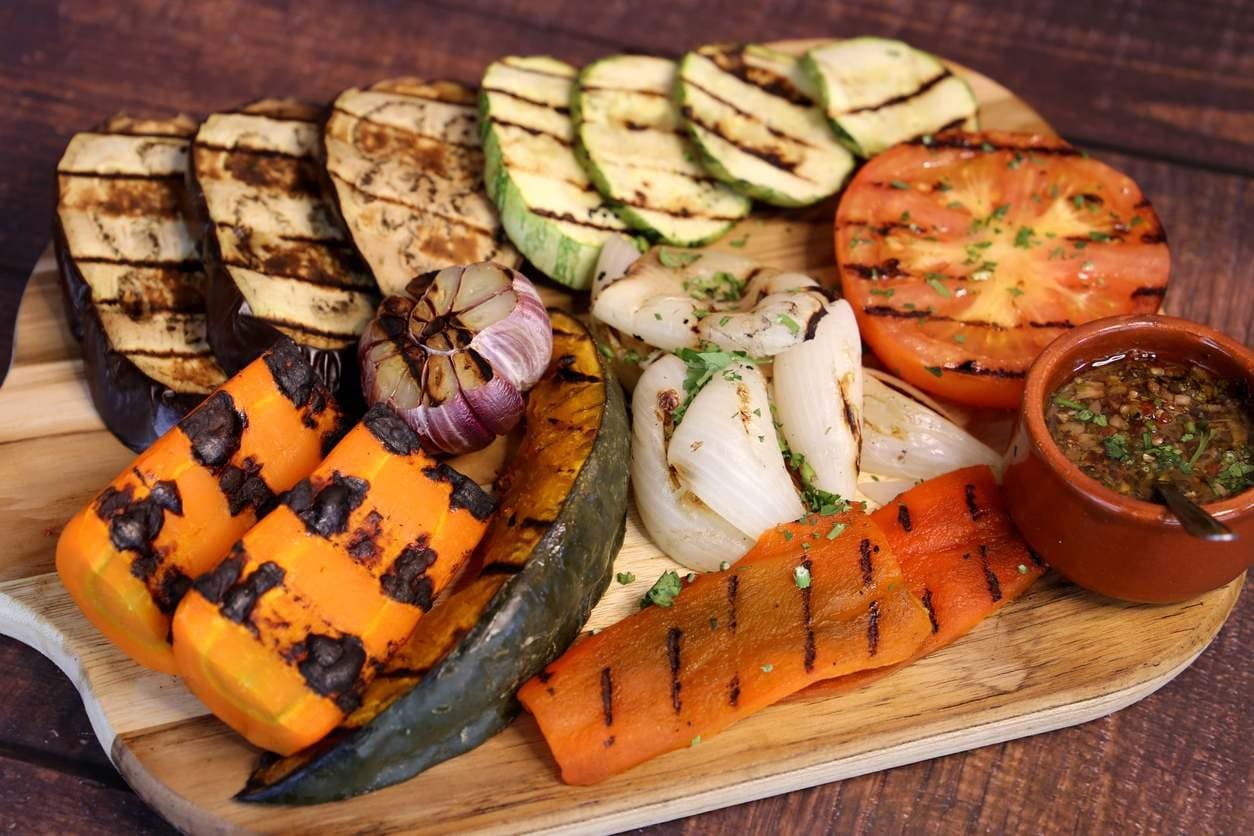 Tábua de madeira com diversos vegetais grelhados como churrasco: abobrinha, berinjela, tomate, abóbora cabotiá, cebola, cenoura e alho.
