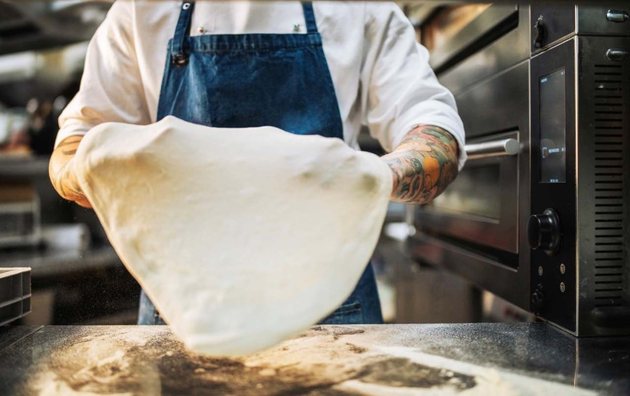 Pizzaiolo abrindo massa de pizza ao lado de forno industrial