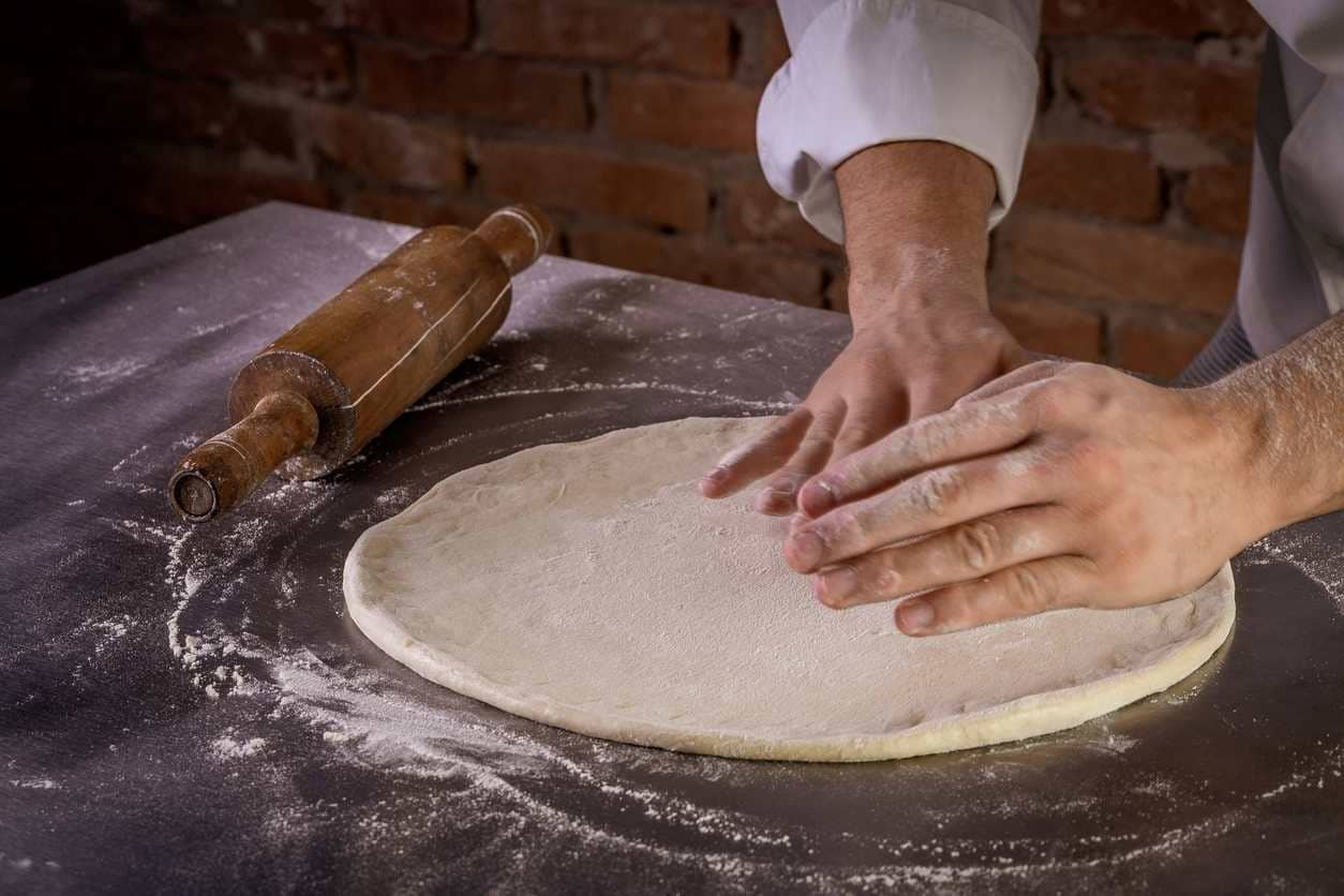 Pizzaiolo abrindo disco de massa de pizza sobre mesa de pedra farinhada, com apoio de rolo de madeira