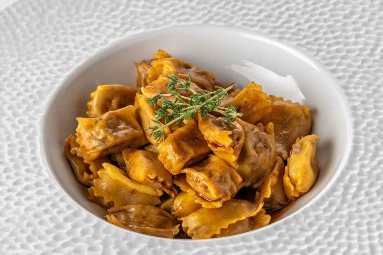 Porção de agnolotti servida em bowl branco, com molho de açafrão e enfeitado com ramo de orégano.
