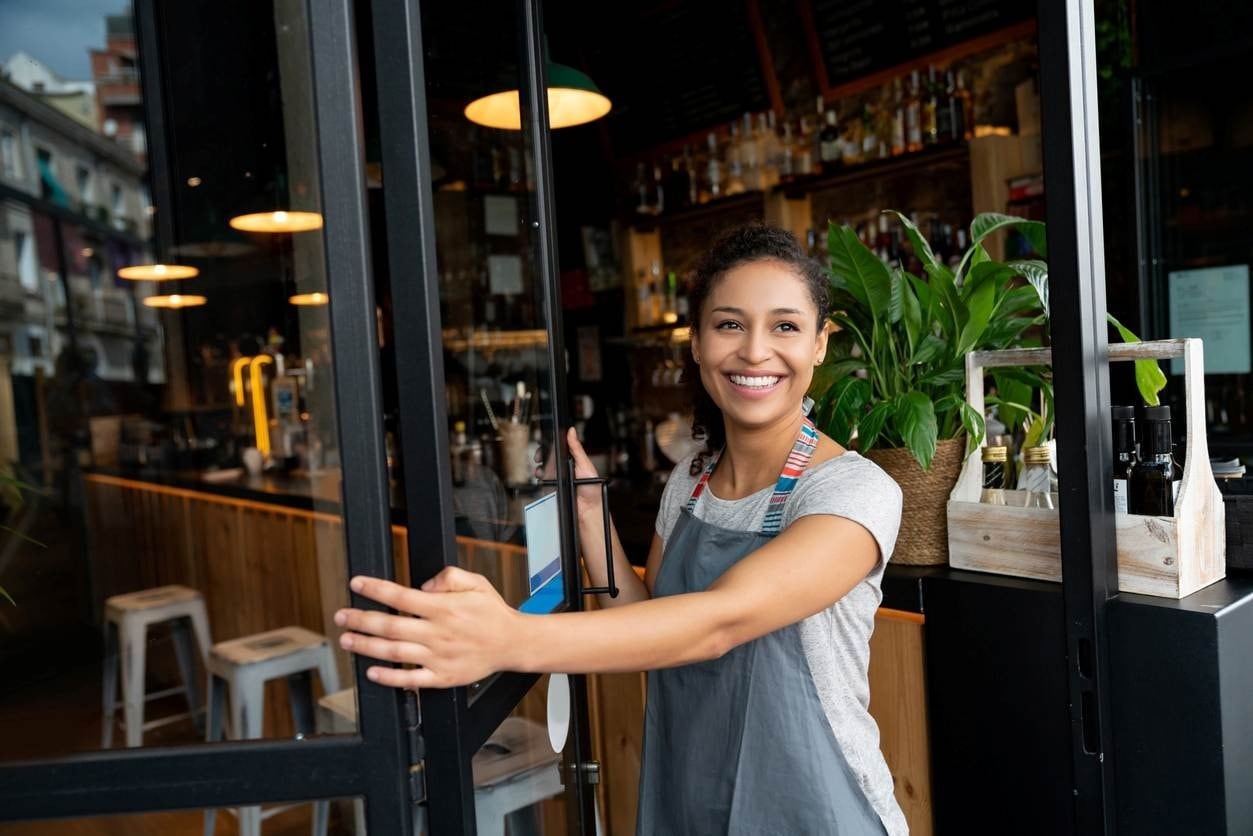 Mulher de avental sorrindo enquanto abre a porta de sua lanchonete, recebendo clientes no estabelecimento.