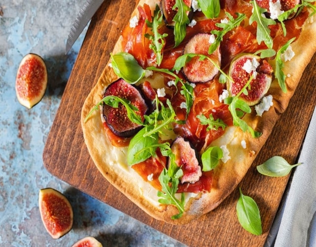 Tábua de madeira com pizza retangular de figo e presunto parma. Ao redor, pedaços de figo e folhas de manjericão. Tábua de madeira com pizza retangular de figo e presunto parma. Ao redor, pedaços de figo e folhas de manjericão.