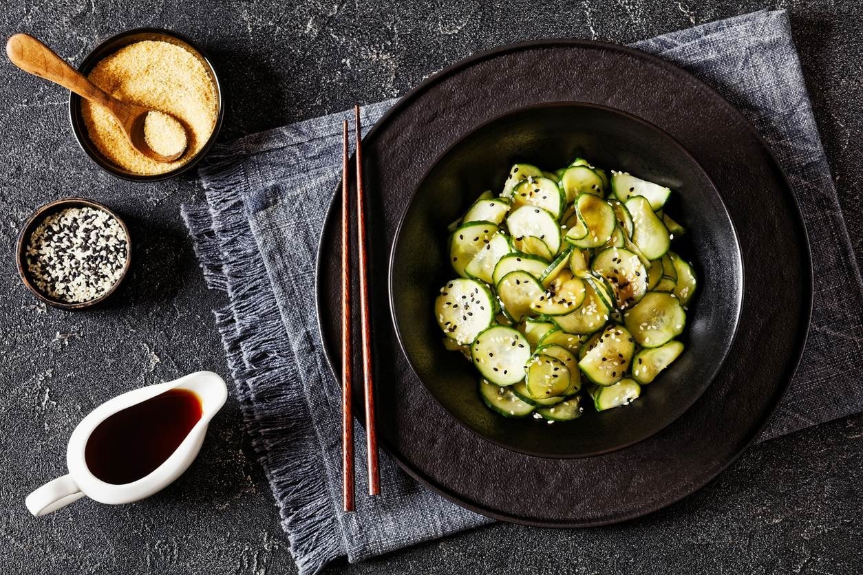 Sunomono de pepino com gergelim em tigela preta, com hashi, shoyu e temperos ao lado.