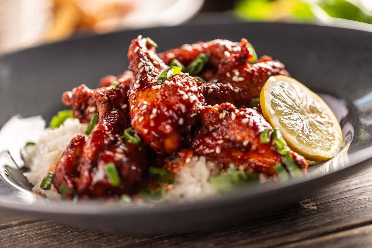 Frango ao molho shoyu com gergelim e cebolinha sobre arroz, com rodela de limão, em prato escuro.