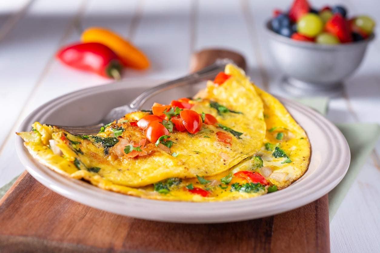 Omelete recheada com tomate, pimentão e ervas servida em prato, com frutas ao fundo.