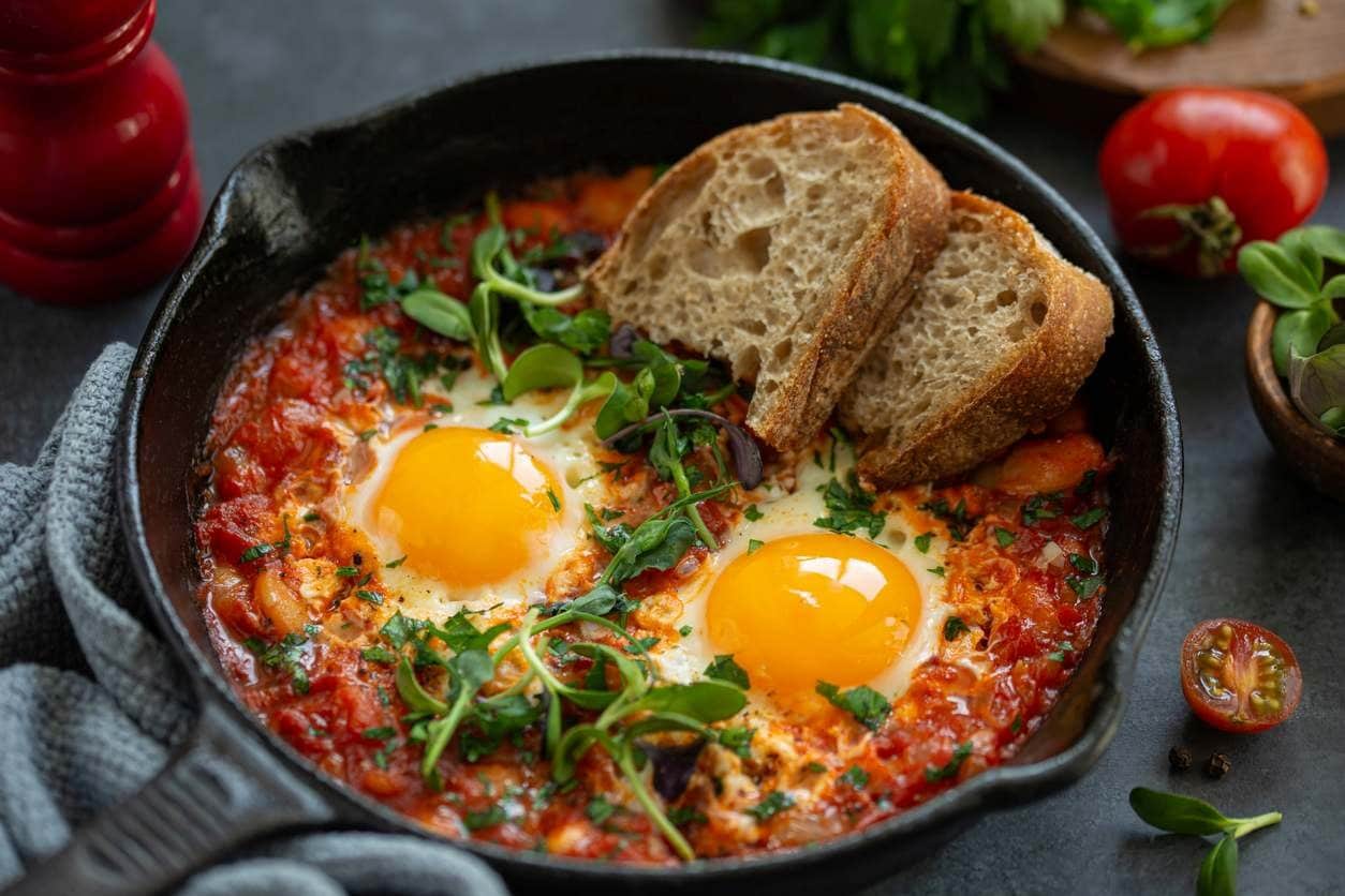 Shakshuka com ovos pochê em molho de tomate, ervas e fatias de pão em frigideira de ferro.