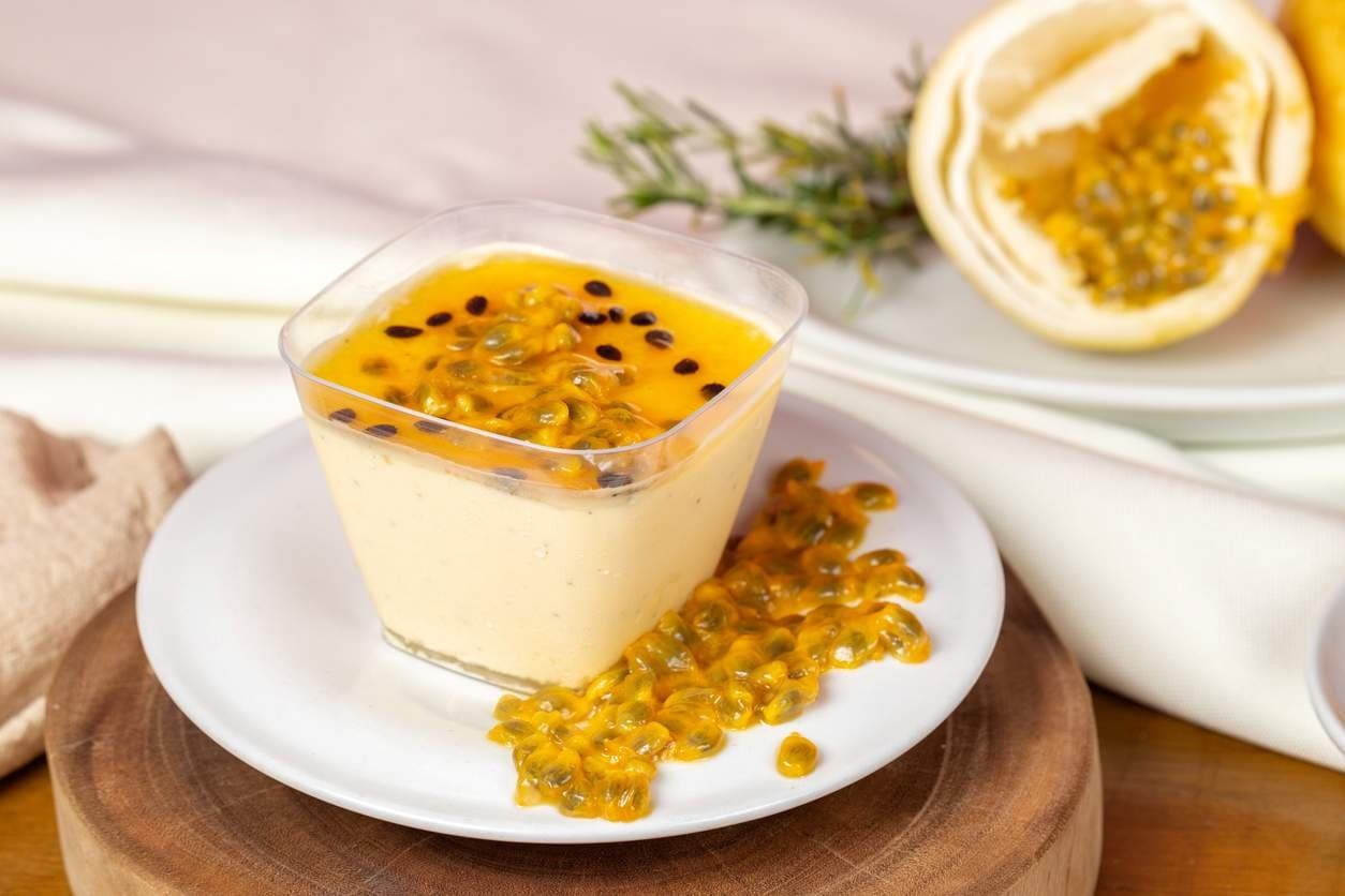 Mousse de maracujá em taça de vidro com calda e sementes da fruta, servida sobre prato branco.