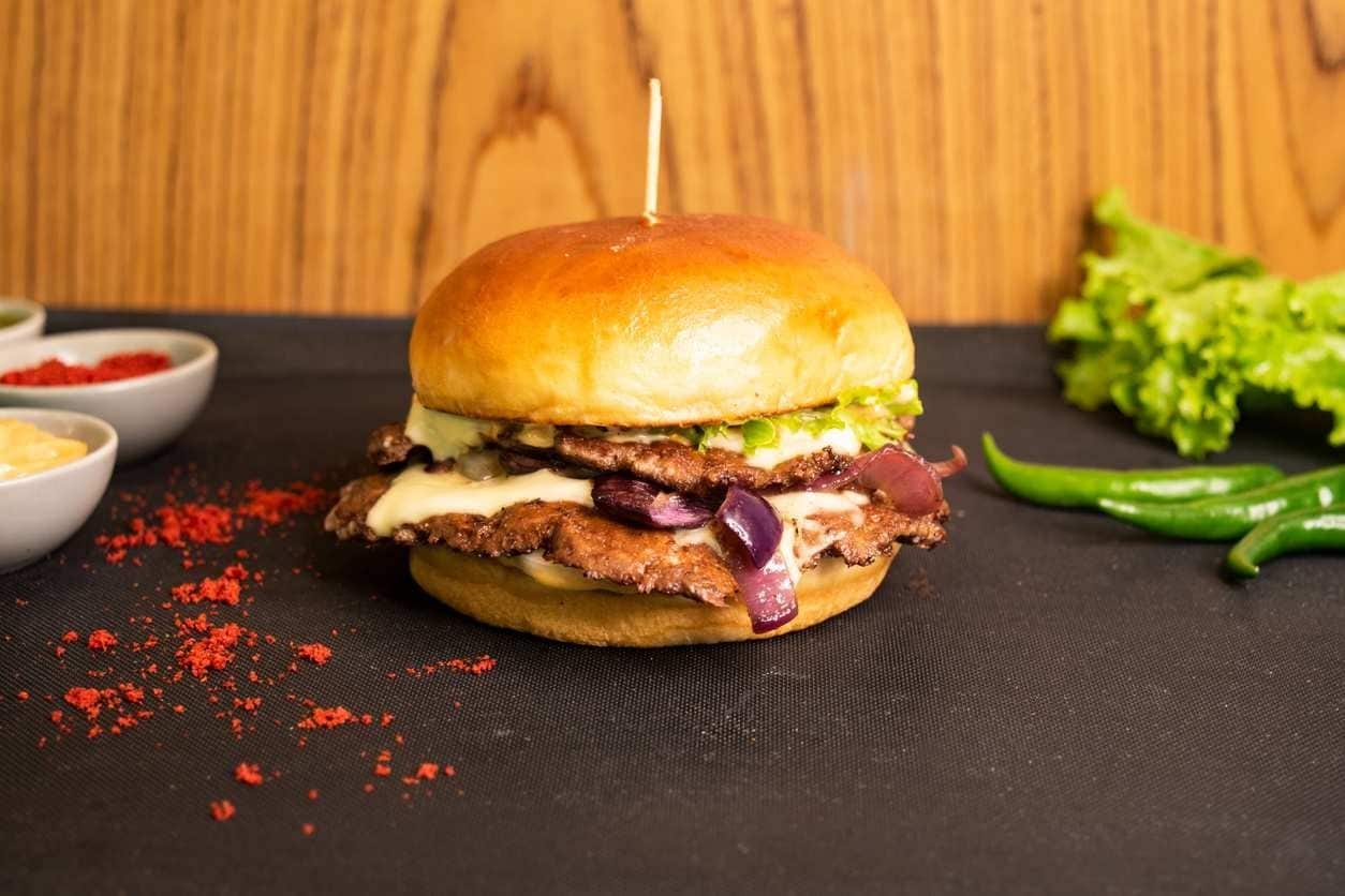 Smash burguer com bacon, cebola caramelizada, picles e salda, em mesa com ingredientes.