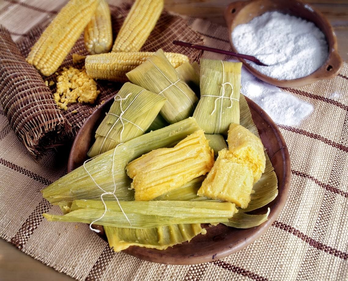 Pamonhas em gamela, algumas já abertas, outras ainda fechadas. Ao redor, espigas e farinha de tapioca compõem a cena. Pamonhas em gamela, algumas já abertas, outras ainda fechadas. Ao redor, espigas e farinha de tapioca compõem a cena.