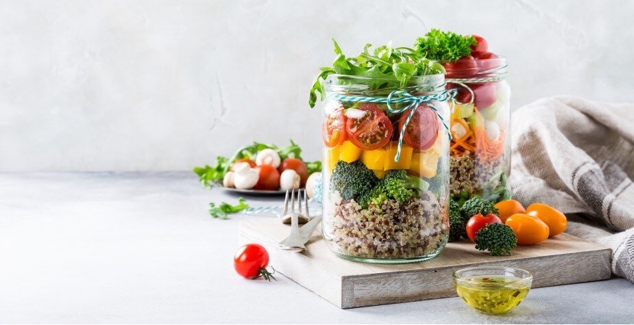 A imagem mostra dois potes de vidro, contendo saladas variadas e coloridas, em um espaço branco que mostra mais ingredientes. A imagem mostra dois potes de vidro, contendo saladas variadas e coloridas, em um espaço branco que mostra mais ingredientes.