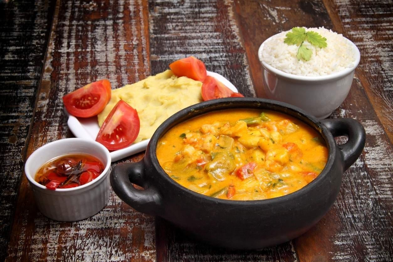 Panela de moqueca de camarão, com arroz, purê de batatas, tomate e pimenta ao redor