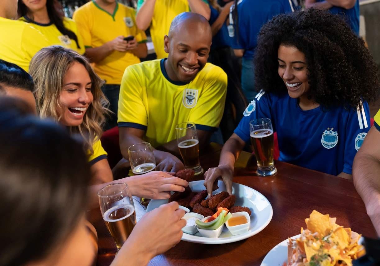 Amigos vestindo camisas de futebol comemoram em bar com cerveja e finger food enquanto assistem a uma partida.