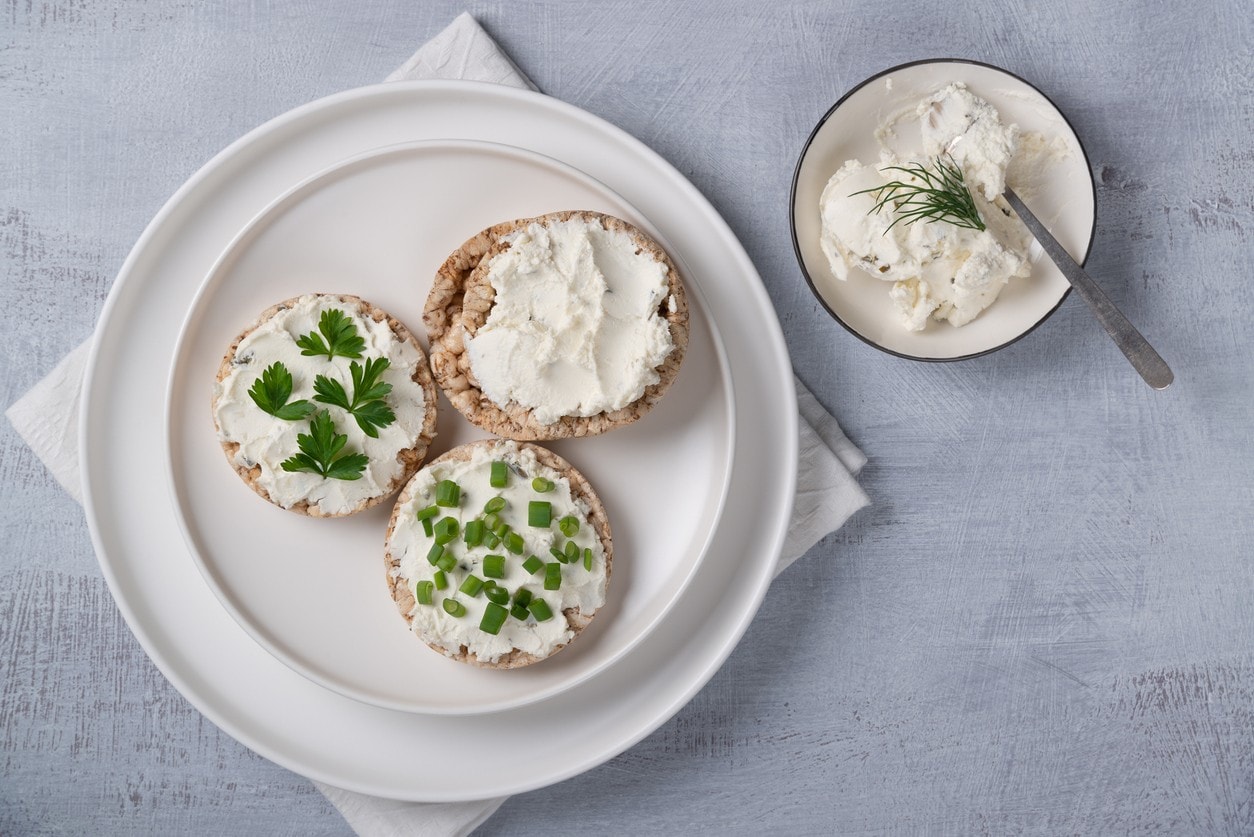 Prato branco contendo três biscoitos de arroz com coalhada seca, um com salsinha, um cebolinha e outro puro. Há uma cumbuca com mais coalhada, com dill por cima. Prato branco contendo três biscoitos de arroz com coalhada seca, um com salsinha, um cebolinha e outro puro. Há uma cumbuca com mais coalhada, com dill por cima.