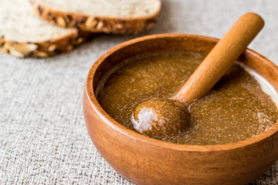 Duas fatias de pão integral ao fundo. No centro, um bowl de madeira com tahine temperado com melaço, pronto para ser servido. Duas fatias de pão integral ao fundo. No centro, um bowl de madeira com tahine temperado com melaço, pronto para ser servido.