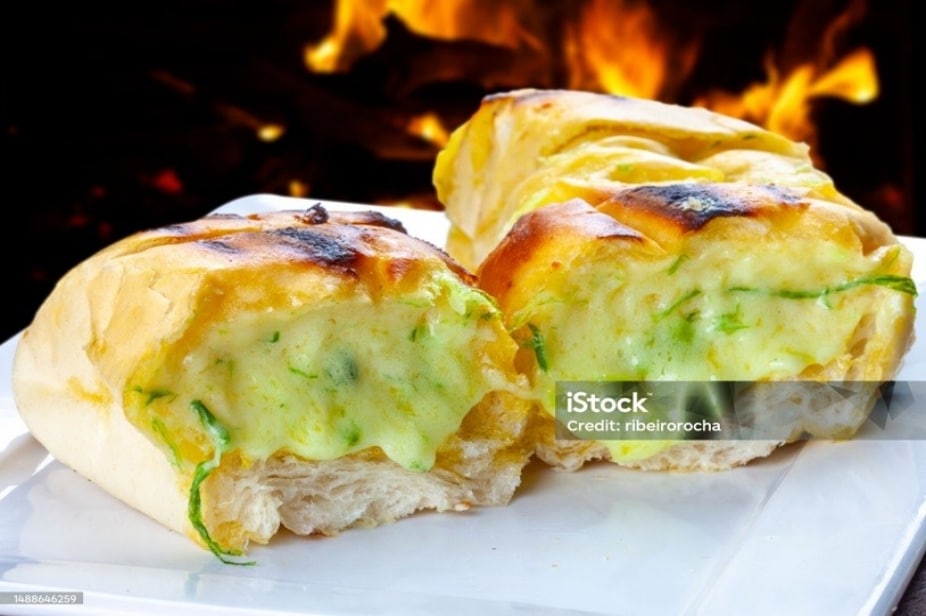 Pão de alho cortado ao meio, recém assado, cremoso e suculento. Ao fundo, vemos a churrasqueira em chamas. Pão de alho cortado ao meio, recém assado, cremoso e suculento. Ao fundo, vemos a churrasqueira em chamas.